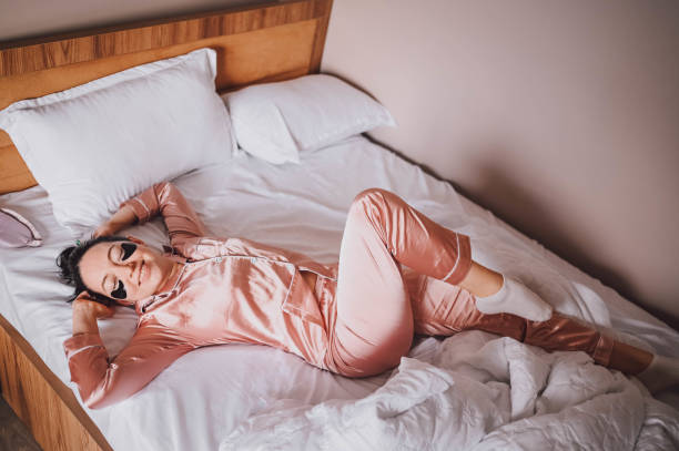 Cosmetology, skin care, face treatment, spa, natural beauty concept. Smiling young woman in pink silk pajamas lies in bed with black moisturizing patches on her eyes. Beauty routine. stock photo