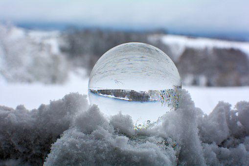 Glass ball lies in the deep snow on a cold winter day in nature, with reflections of the winter landscape in the glass