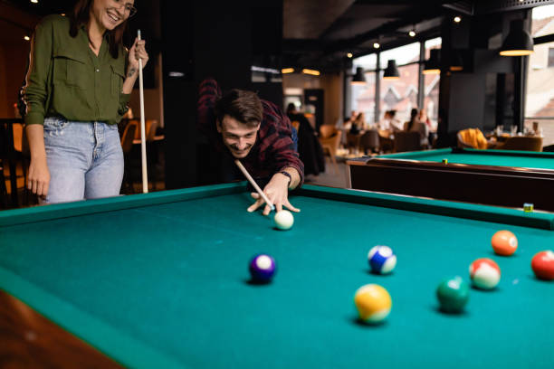 una giovane coppia di innamorati è in competizione su chi metterà più palle da biliardo nelle buche. ci sono persone sullo sfondo. - pool game pool table aiming men foto e immagini stock