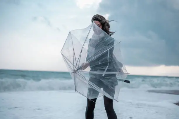 Photo of Sad young woman walking alone on the beach with umbrella in rainy weather