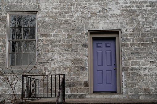 Facade of a stone building with a freshly painted purple front door and a window.  A reflection of bare trees in winter is reflected in the window pane.