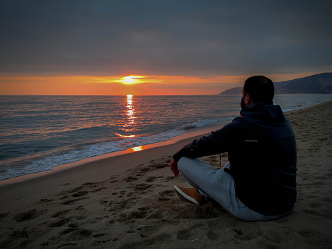 Young man sitting on the beach regarding beautiful sunset over mediterranean sea at autumn