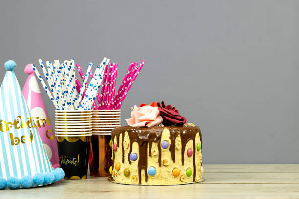 istock Birthday layer cake decorated with chocolate pieces and happy birthday text, hazelnuts and roses flowers and disposable cups and straws on wood table 1360845122