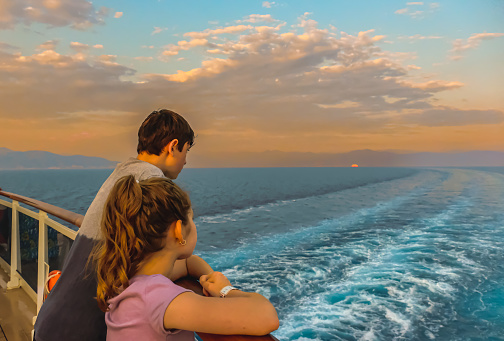Teenage boy and his pre-teen sister watching the wake from in the stern of the ship at sunset