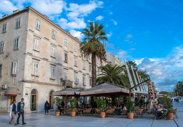 The Riva, waterfront walkway lined with restaurants and cafes, a popular gathering place for locals and tourists in Split, Croatia. Awnings cover outdoor tables and chairs where people enjoy drinks and meals. Clear day with blue skies filled with puffy white clouds. A snapshot of Croatia lifestyle.
