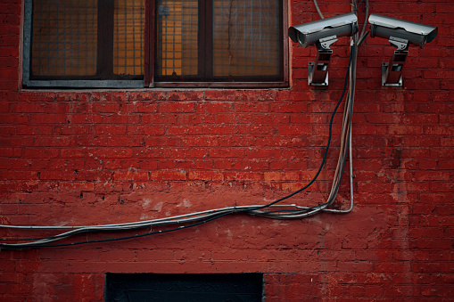 Two metallic surveillance cameras on red brick wall with barred window. Electricity cables on the wall.
