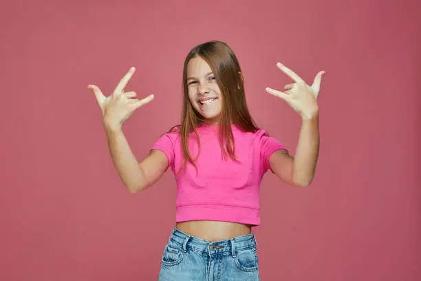 Cool naughty child girl tomboy show rock, heavy metal gesture by hands, having fun on pink studio background. High quality photo