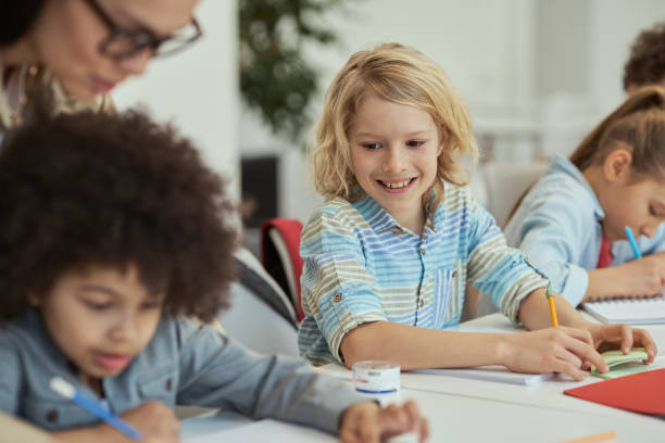 istock Active little schoolboy smiling and looking at his classmate while sitting at the table in elementary school classroom together with other kids 1360857700