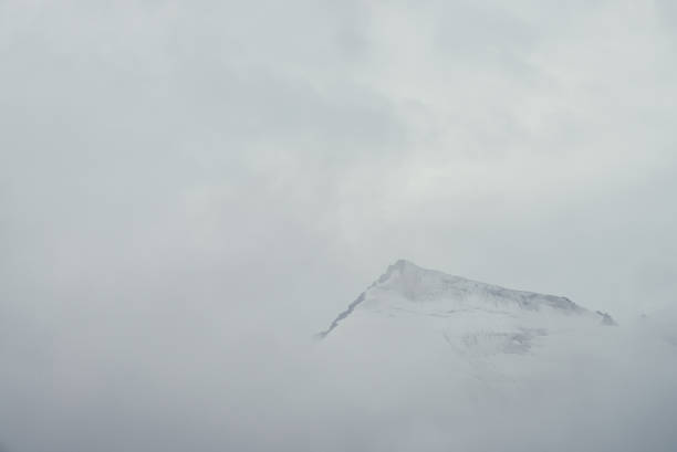 istock Minimal mountain landscape with high pointy rock in clouds. Minimalist mountain scenery with sharp snowy mountain peak over clouds. Snow-white pointed pinnacle above white clouds. Big top in dense fog 1360866657