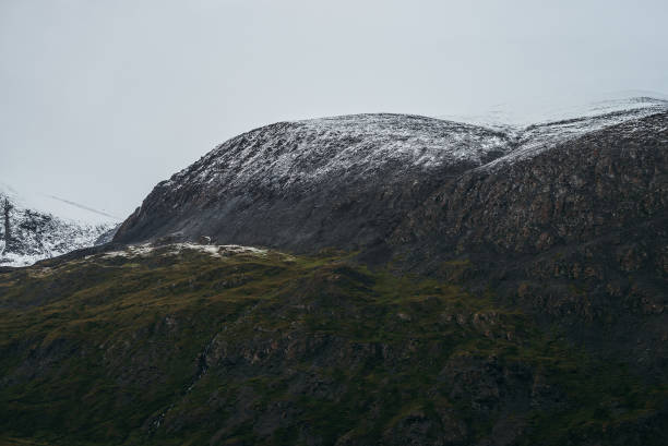 istock Minimalist highlands landscape with snow-capped mountains under cloudy sky. Snowbound high mountain range in overcast weather. Atmospheric minimalism with green mountains, white snow and gray sky. 1360866661