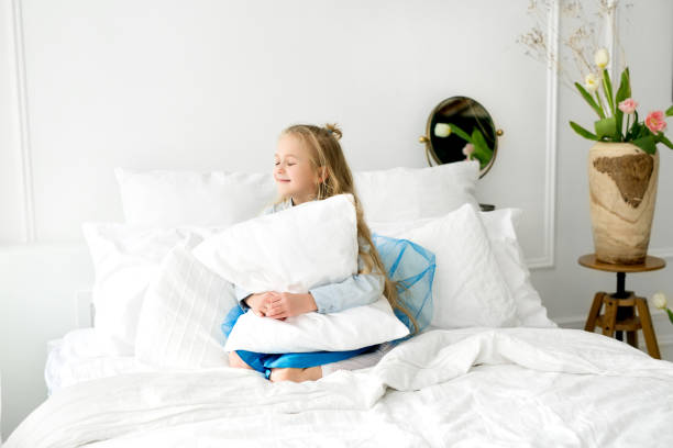 menina está sentada em uma cama com roupa de cama branca de neve, abraçando um travesseiro - baby cheerful child blanket - fotografias e filmes do acervo