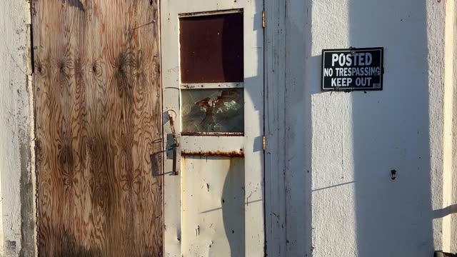 Obsolete building with boarded windows and shattered glass screen door with no trespassing sign posted