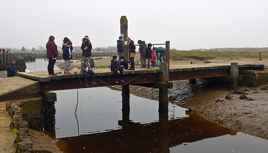 A full-length shot of a father, wife, and two daughters enjoying the view from a pier going into the sea. They are all wearing warm clothing on a cold morning. Located in Amble, Northumberland.