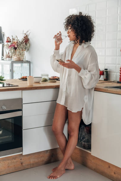 istock Young African American woman drinking water and using cellphone in the kitchen. Vertical image 1360904982