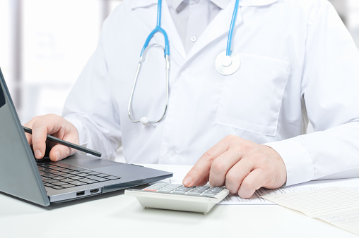 Doctor working with medical statistics and financial reports. doctor counts on calculator and enters data into laptop. doctor working with laptop and calculator researching some medication information