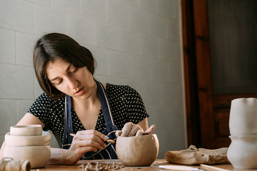 An Armenian girl makes a clay product. Modeling in a home workshop. Creativity and handmade. Clay pot in the hands of a woman with a tool. Art and texture concept.