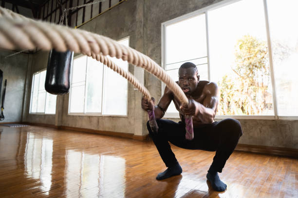 istock African sport man doing battle ropes functional training at gym. Determined trainer making waves with ropes while exercising strength. Athlete working out with battle rope at industrial gym. 1360910051