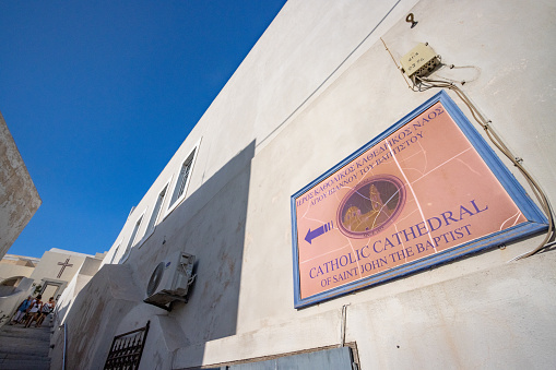 Sign to Catholic Cathedral of St John the Baptist in Firá on Santorini in South Aegean Islands, Greece, with identifiable details visible.