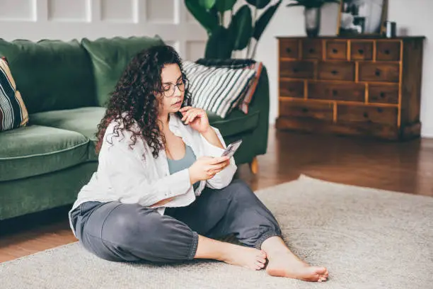 Curly haired plus size young woman wearing comfortable clothes in glasses clicks on smartphone screen sitting on floor mat in stylish room