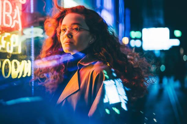 Curly haired young woman tourist with light makeup in glasses looks around standing near bar with colorful neon sign against night megalopolis stock photo