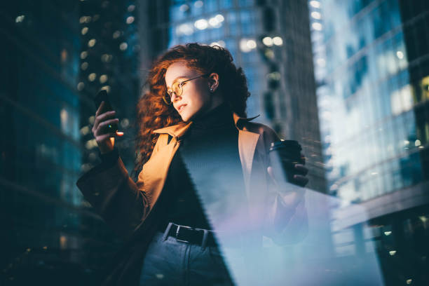 Successful woman using smartphone outdoors while standing near skyscraper at night. stock photo