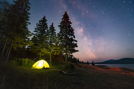 Milky Way and sky full of stars above the luminous tent under the trees by the lake