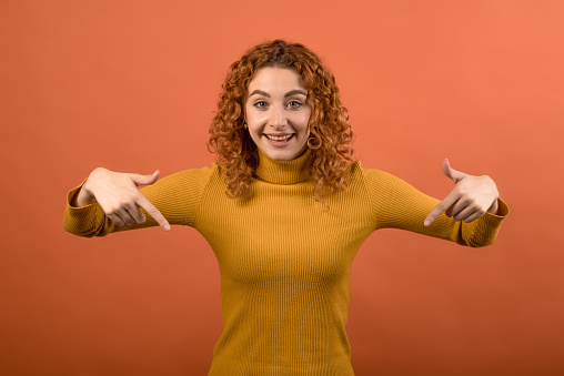 Young and attractive Caucasian redhead girl in orange jumper pointing down at copy space isolated on orange studio background.