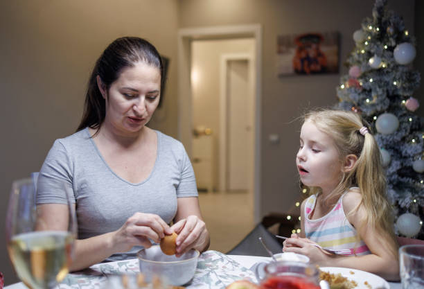 little blond girl is watching her mom who is shelling a boiled egg at the dinner table in the room in which there is a christmas tree. - drinking little girls women wine imagens e fotografias de stock