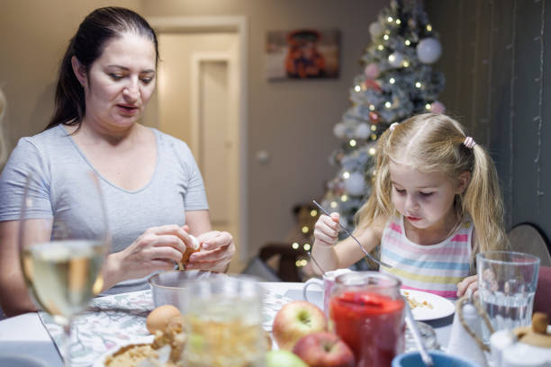 little girl with long blond hair and her mother are eating at a table in the room in which there is a christmas tree. - drinking little girls women wine imagens e fotografias de stock