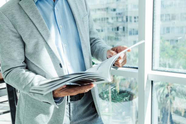 hombre de negocios en traje de negocios lee una revista cerca de la ventana con una planta - folleto fotografías e imágenes de stock