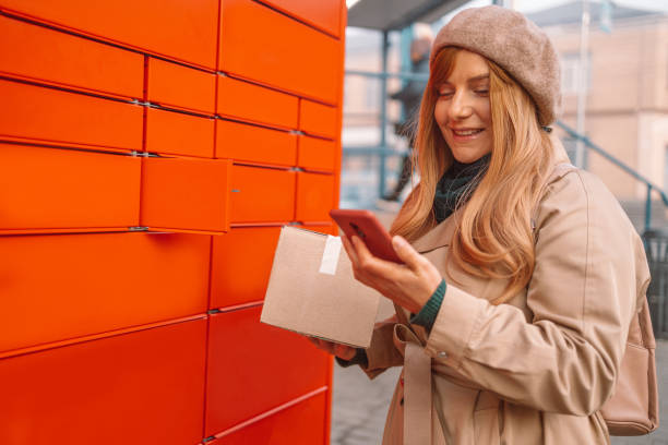 istock Young caucasian woman holding smartphone using automated self service post terminal machine or locker to deposit the parcel for storage 1361026970