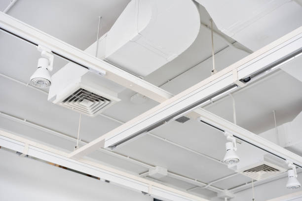 Ceiling mounted cassette type air condition units with other parts of ventilation system (tubes, cables and vents) located inside commercial hall with hanging lights and other construction parts.
