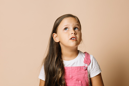 Portrait of child with shining bright eyes looking upward and eyebrows raised, with pensive, curious look wearing bright pink jumpsuit and white t-shirt on beige background.
