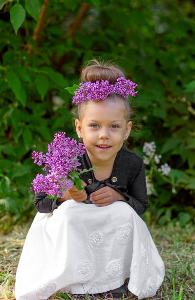 tamanho de corpo de comprimento completo sorrindo criança branca de 5 anos com coroa lilás na cabeça e buquê lilás nas mãos olhando para a câmera agachada em parque verde no fundo de árvores - child 4 5 years laughing little girls - fotografias e filmes do acervo