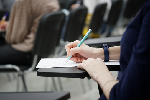 A girl writes a dictation or fills out documents in the audience, sitting on a school chair with a writing stand. Close-up. No face