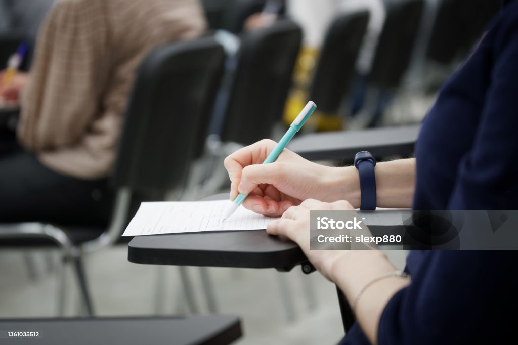 A girl writes a dictation or fills out documents in the audience, sitting on a school chair with a writing stand. Close-up A girl writes a dictation or fills out documents in the audience, sitting on a school chair with a writing stand. Close-up. No face Educational Exam Stock Photo