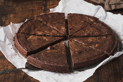 Sticky chocolate brownie cake, mud cake or traditional swedish kladdkaka, sliced into eight pieces. The cake is on a parchment paper, on a rustic old wooden table.