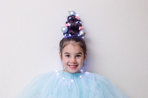 Portrait of funny beautiful girl with a hairstyle in the form of a Christmas tree with a luminous garland, stands against the background of a light wall