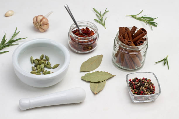 Cinnamon sticks in jar. Cardamom in mortar and pestle on table. Allspice in glass bowl. Paprika petals in jar. Bay leaf and rosemary on table. Top view. White background.