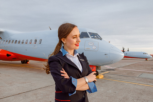 Joyful female flight attendant keeping arms crossed and smiling while standing at airfield with passenger airplane on background