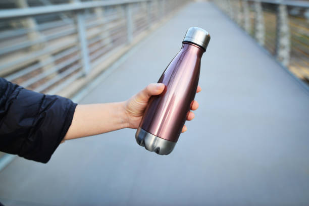 primer plano de la mano femenina sosteniendo una botella termo brillante de acero reutilizable para agua. concepto de espacio de copia. botella de acero para agua en el carril bici. - water bottle bottle steel stainless steel fotografías e imágenes de stock