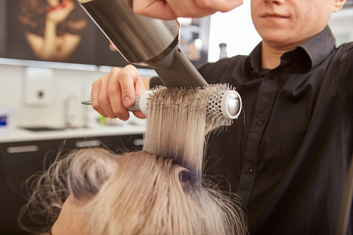 Close up of male hairstylist using hairdryer and round brush while styling young woman hair