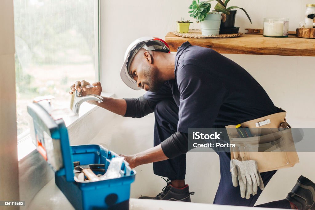 Plumber fixing a leaking bathroom faucet African male plumber looking at leaking bathroom tap. Professional handyman trying to fix a bathroom faucet. Plumber Stock Photo
