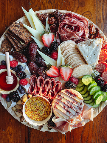 Charcuterie Board or Tray piled high with savory delights such as honey, rye crackers, dates, strawberries, pistachio nuts, walnuts, blueberries, raspberries, mustard, cheeses, cucumbers and Italian meats. View from directly above.