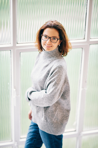 Interior portrait of beautiful 40 year old woman relaxing next to window, wearing warm grey pullover and glasses
