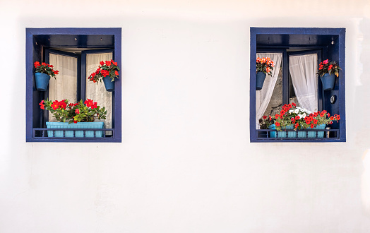 two blue windows decorated with flower pots with red flowers on a house with white walls, typical mediterranean village, copy space horizontal