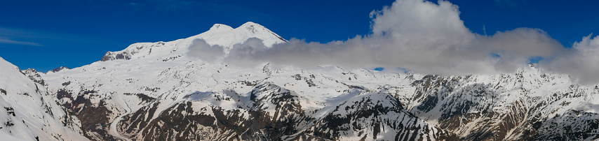 Panoramic view of Mount Elbrus with snow-capped peaks against the blue sky. Elbrus is a 5642 meter high stratovolcano located in Karbardino-Balkaria, Russia.