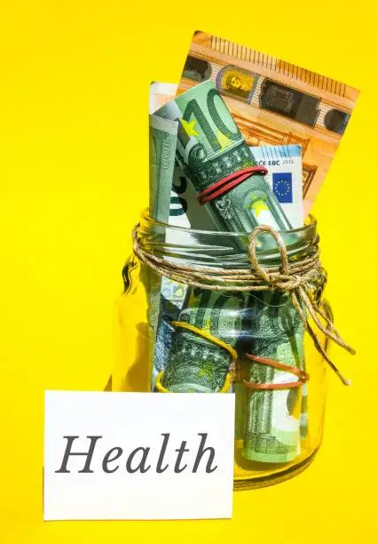 Photo of Glass jars filled with Euro bills, savings inside glass jar, money isolated on yellow background. Paper note written word HEALTH. Business budget of wealth