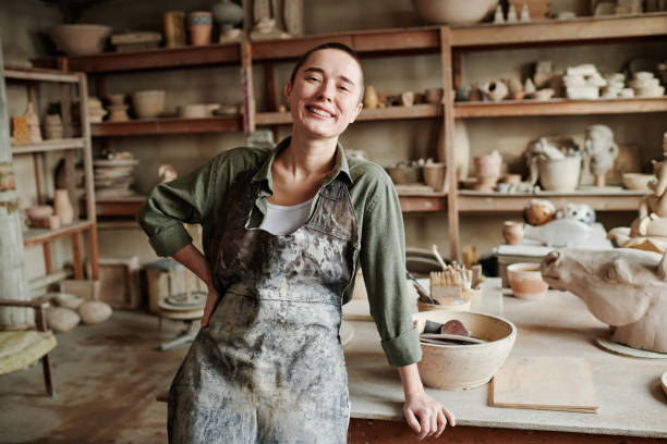 Successful scultor working in workshop Portrait of young happy woman in apron smiling at camera while working in ceramic workshop pottery making stock pictures, royalty-free photos & images
