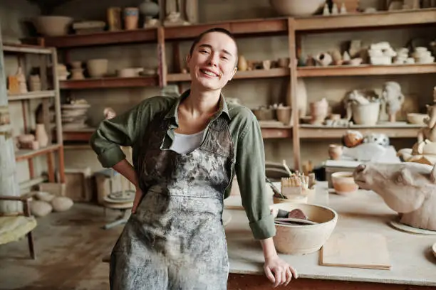Portrait of young happy woman in apron smiling at camera while working in ceramic workshop